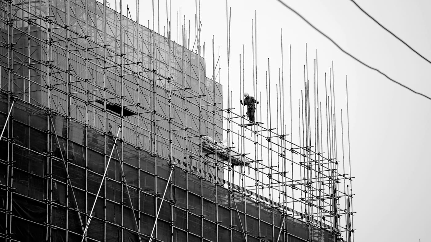 防護シートで覆われた建物外周の足場と、上部で作業する職人（サンプル写真）